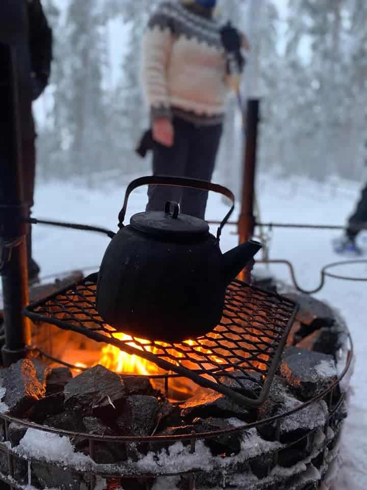 Kodassa on tulet ja porukka istuu ympärillä. Pyörätuolissa oleva mies on lisäämässä puita pesään.