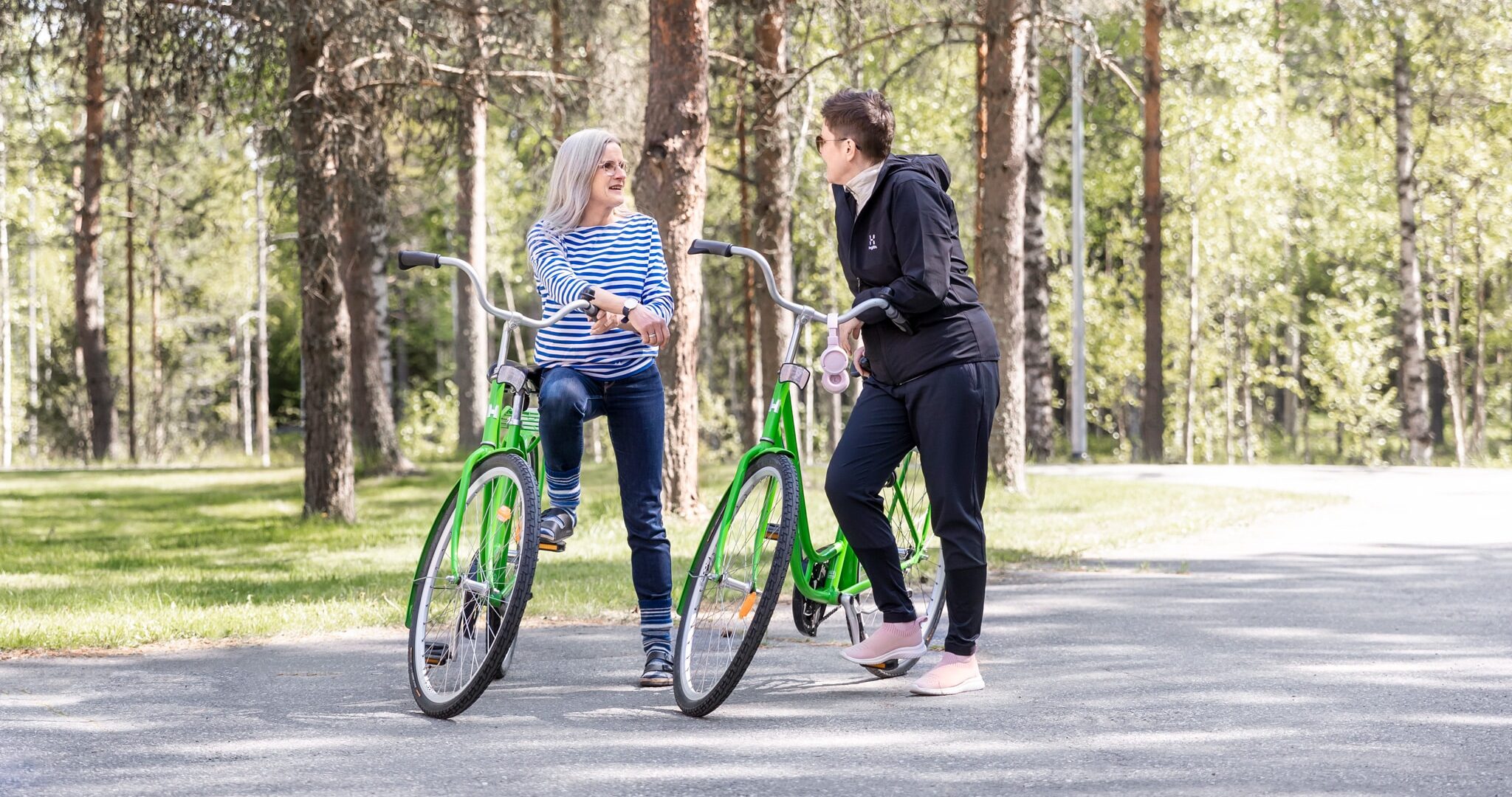 Kaksi naista nojailee vihreisiin polkupyöriin asfalttipolulla. Takana on metsää.