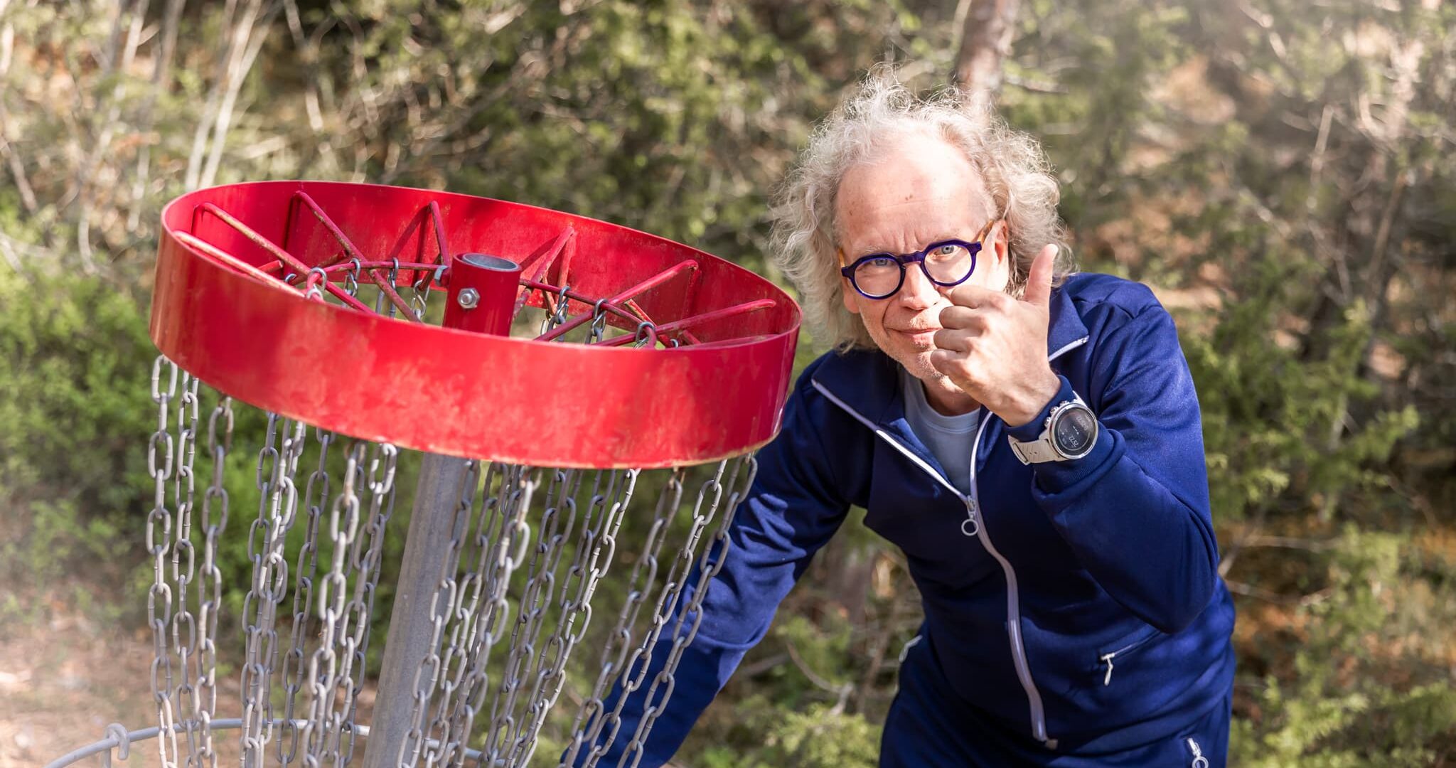 Mies näyttää peukkua frisbeegolf-korin vieressä. Taustalla vihreää kangasmetsää.