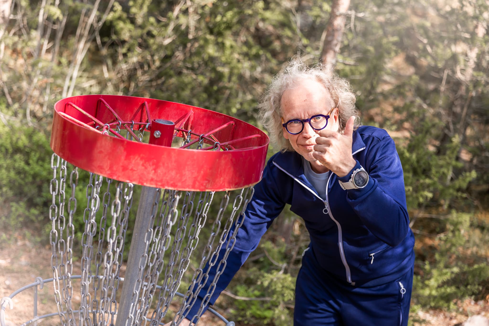 Mies näyttää peukkua frisbeegolf-korin vieressä. Taustalla vihreää kangasmetsää.