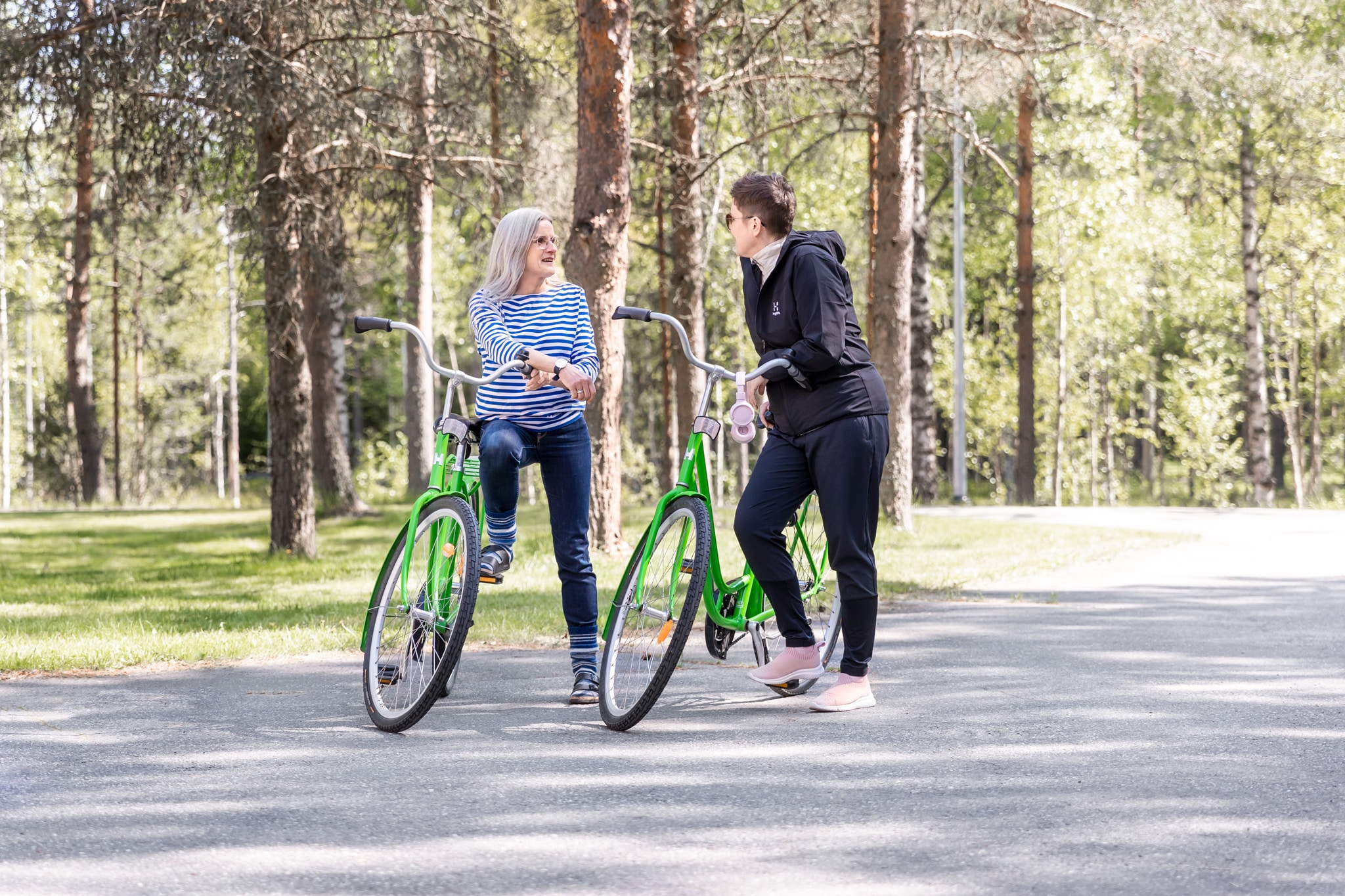 Kaksi naista juttelee ja nojailee vihreisiin pyöriin. Taustalla keväistä mäntymetsää.