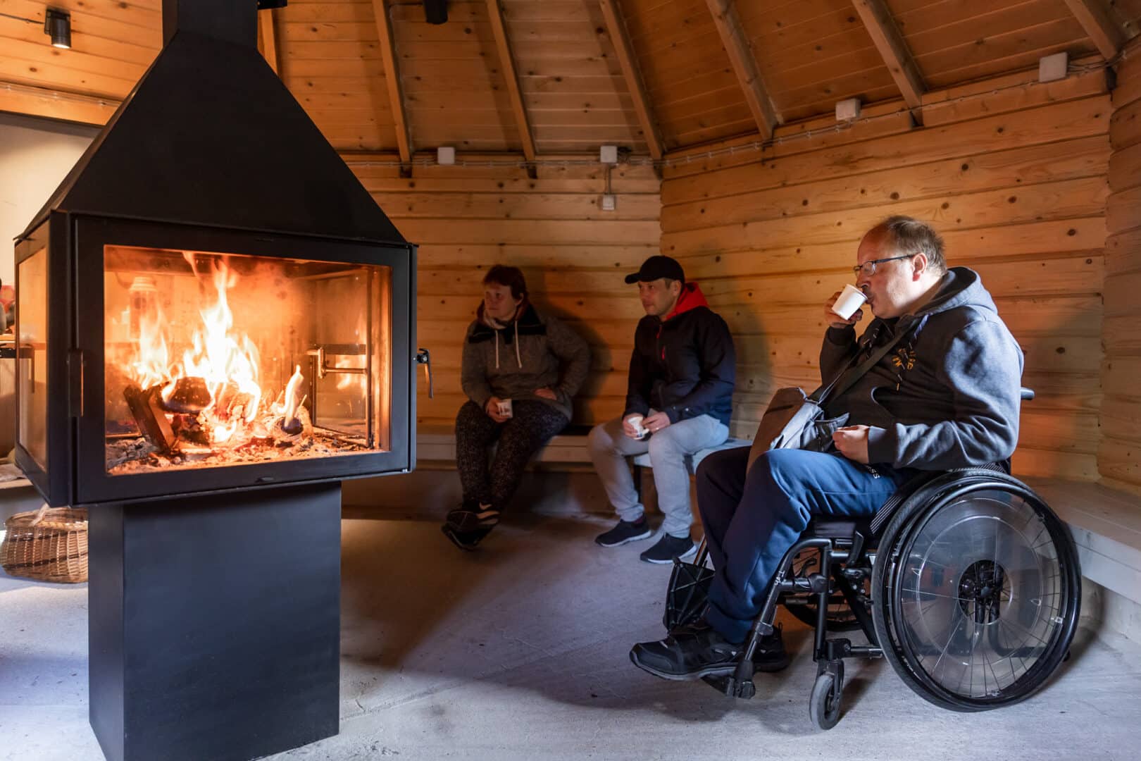 Kuva esteettömästä kodasta, jossa on tulet. Etualalla pyörätuolissa oleva mies juo kahvia. Taustalla Nainen ja mies istuvat seinustan penkillä.