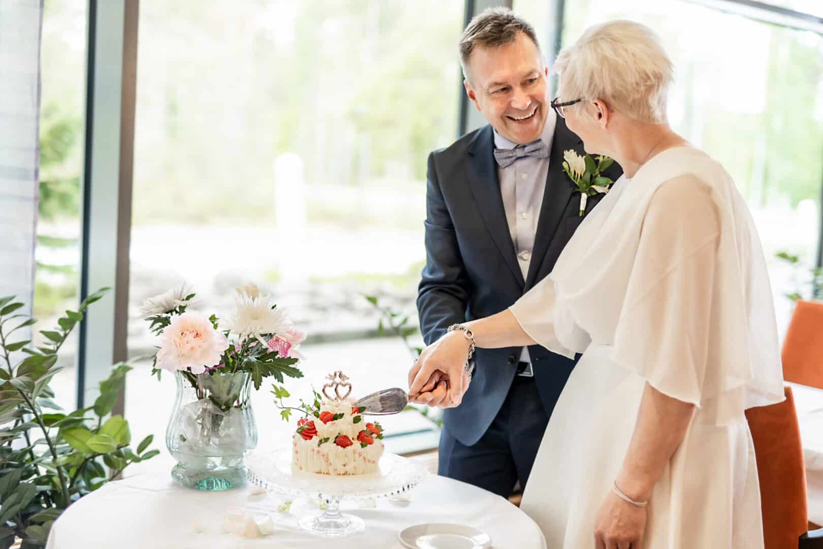 Hääpari leikkaa hääkakkua ravintolasalin isojen ikkunoiden edessä. Katsovat toisiaan silmiin ja hymyilevät.