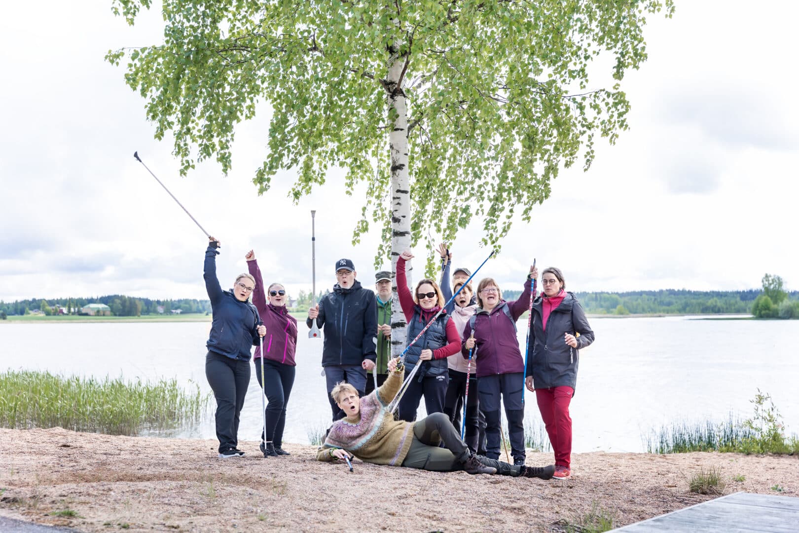 Kuntoutusporukka koivun alla iloisina kävelysauvat ilmassa. Taustalla järvimaisema.