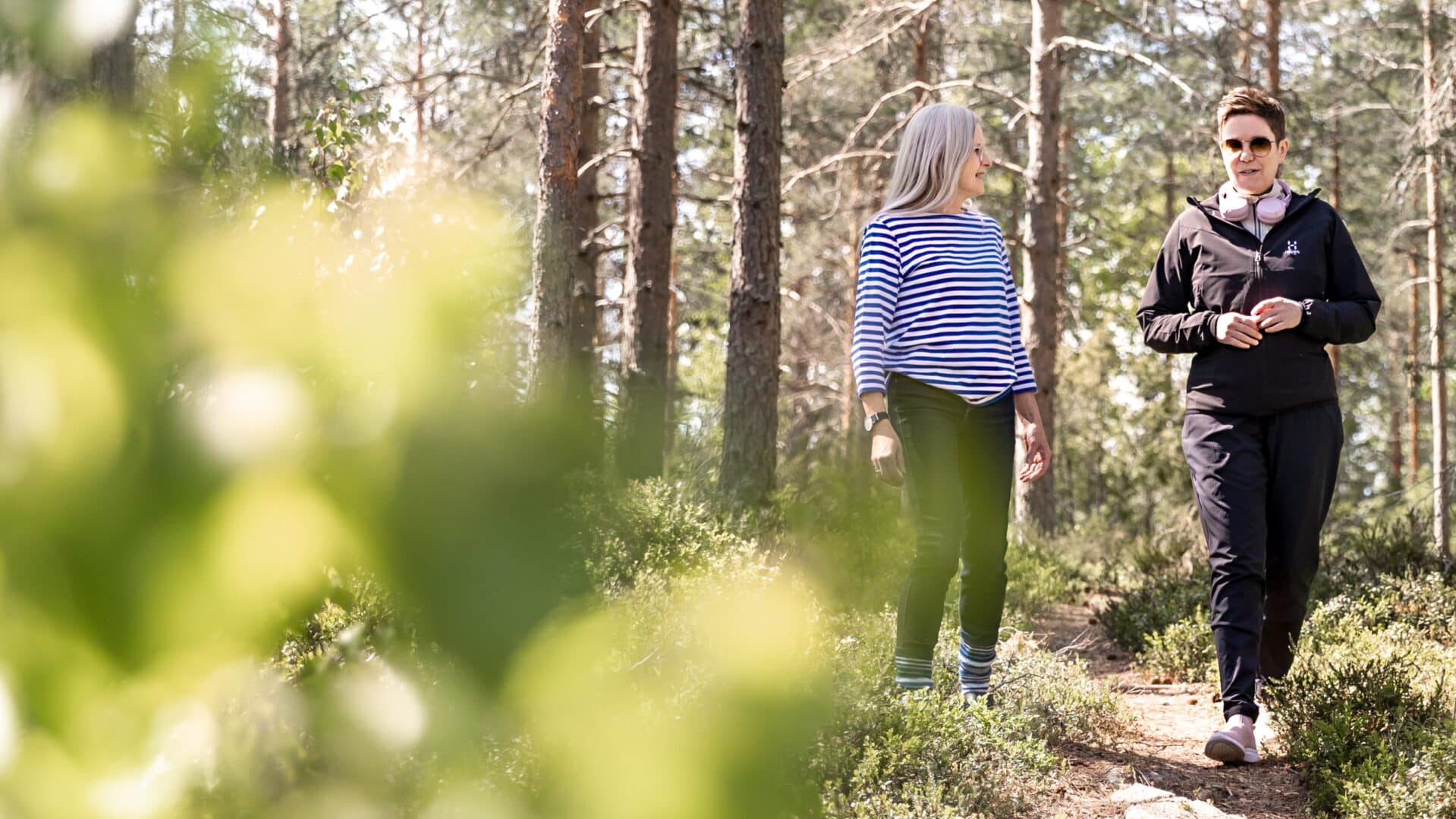 Kaksi naista kävelee polulla keväisessä mäntymetsässä kameraa kohti