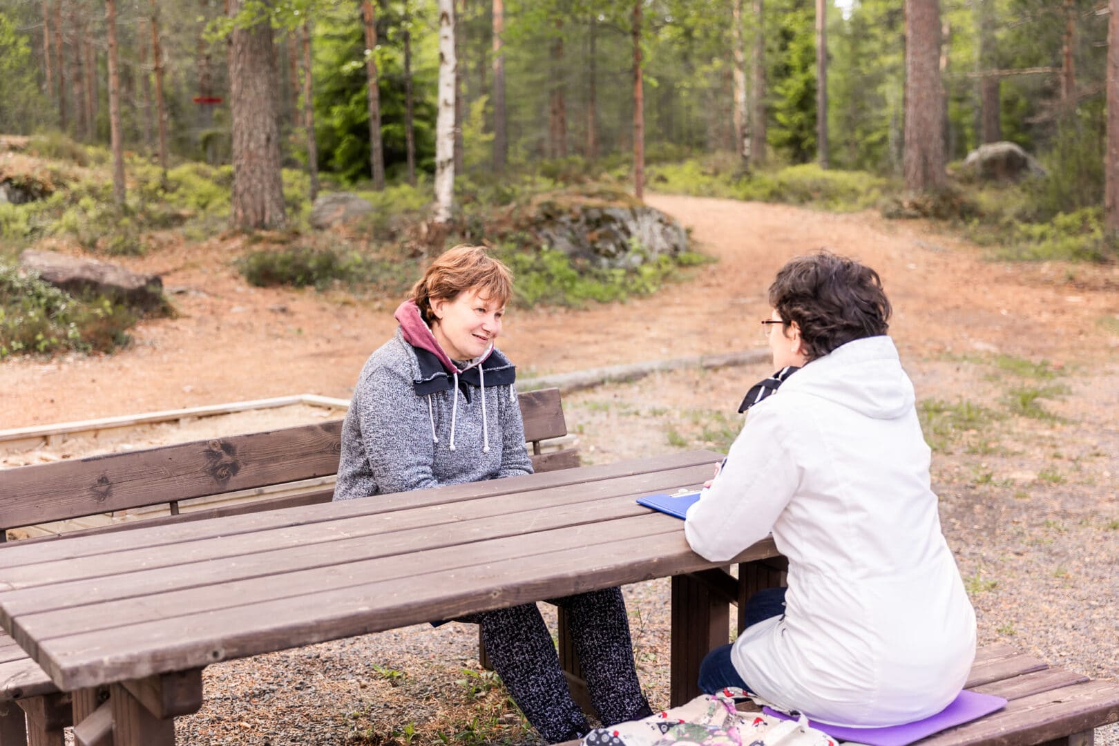 Moniammatillisessa kuntoutuksessa sairaanhoitaja ja asiakas juttelevat ulkona pöydän ääressä. Takana on metsää ja pururata.